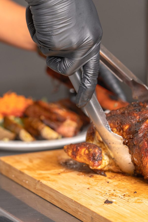 Gloved hands using tongs to serve a whole grilled chicken on a wooden cutting board in a commercial kitchen